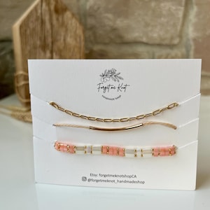 Może przedstawiać: Zestaw trzech delikatnych bransoletek na białej karcie. Górna bransoletka to złoty łańcuszek, środkowa ma złoty pręt, a dolna ma różowe i białe koraliki. Karta zawiera nazwę sklepu i dane social media.