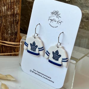 May include: White and blue hockey jersey earrings with the text "TORONTO MAPLE LEAFS". The earrings are displayed on a white card with the text "Forget me Knot Handmade Shop".