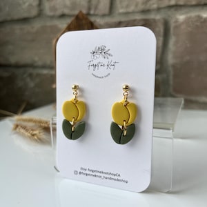 Op de afbeelding: Handgemaakte oorbellen in geel en olijfgroen, gepresenteerd op een witte kaart. De kaart bevat de tekst "Forget Me Knot Handmade Shop". De oorbellen hebben goudkleurige pinnen. Een eigentijds ontwerp.