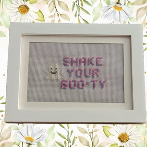 May include: A white framed cross-stitch with a smiling yellow sun and the text "Shake Your Booty" in pink.