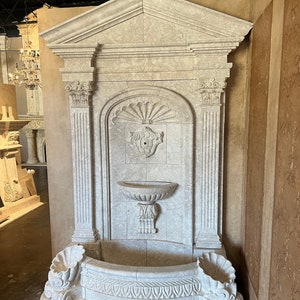 May include: A white stone fountain with a decorative shell design. The fountain has a rectangular basin with a curved front and a small, round basin at the top. The fountain is made of a light-colored stone with a textured surface.