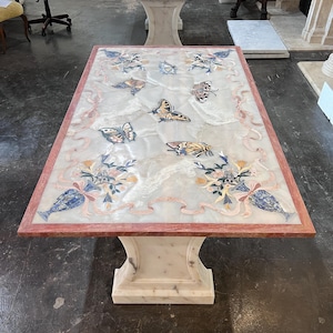 May include: Rectangular marble table with a white surface, adorned with butterfly and floral mosaic designs. The table has a red border and a white pedestal base. A second table is visible in the background.