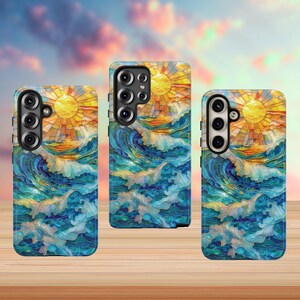 May include: Three phone cases with a stained-glass design featuring a yellow sun, blue ocean waves, and an orange horizon. The cases are on a wooden surface with a blurred sky background.