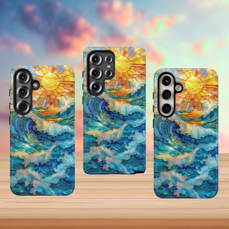 May include: Three phone cases with a stained-glass design featuring a yellow sun, blue ocean waves, and an orange horizon. The cases are on a wooden surface with a blurred sky background.