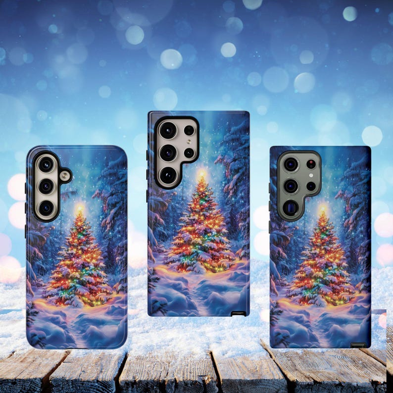 May include: Three phone cases with a winter scene featuring a decorated Christmas tree. The cases have a blue background with a snowy forest and a brightly lit tree. The cases are on a wooden surface.