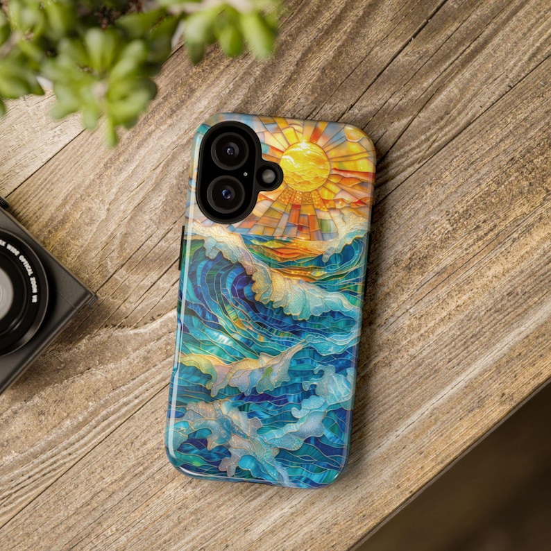 May include: A colorful phone case featuring a mosaic design of a sun over ocean waves. The artwork uses shades of blue, yellow, and orange. The case is on a wooden surface.