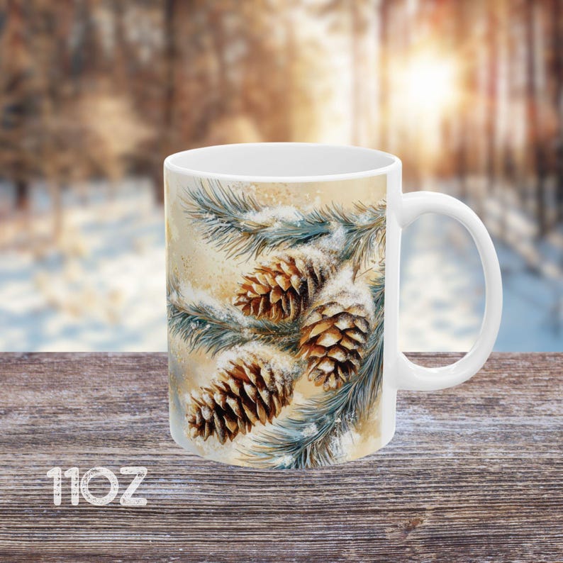 Peut inclure: Une tasse en c&eacute;ramique blanche avec une sc&egrave;ne d'hiver repr&eacute;sentant des branches de pin avec de la neige et des pommes de pin. Les pommes de pin sont brunes et les branches sont vertes.