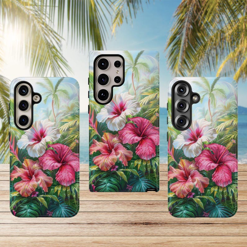 May include: Three phone cases with a tropical floral design. The cases feature vibrant pink and white hibiscus flowers, lush green foliage, and palm trees against a blue sky and ocean backdrop. The cases are displayed on a wooden surface.