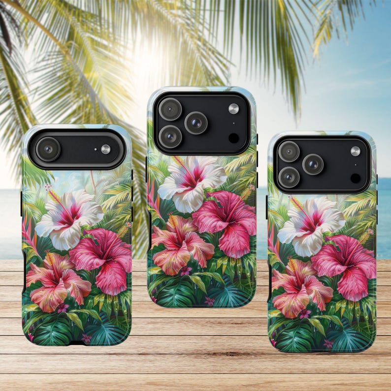 May include: Three phone cases with a tropical floral design featuring hibiscus flowers in shades of pink and white, set against a backdrop of lush green foliage. The cases are displayed on a wooden surface with a blurred background of palm trees and ocean.