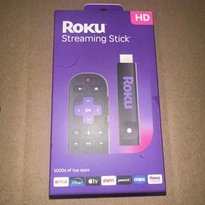 May include: A purple Roku Streaming Stick box with a remote control and the streaming stick itself. The box has the Roku logo and the words "Streaming Stick" printed on it. The remote control has a directional pad and several buttons. The streaming stick is black with the Roku logo.