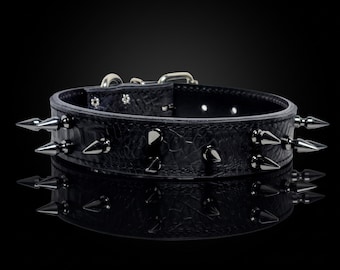 Puede incluir: Un collar negro texturizado que presenta dos filas de púas metálicas afiladas en tono plateado. El collar incluye una hebilla de metal plateado y una anilla en D en la parte superior, con una fila visible de agujeros de ajuste.