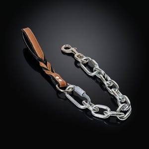 May include: A pet leash featuring a braided tan and black leather handle with two silver-tone rivets, connected to a heavy-duty silver-tone metal chain link section. The chain includes a secure screw-lock carabiner and a swivel snap hook attachment.