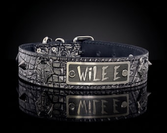 Puede incluir: Un collar de cuero negro con un patrón de cocodrilo texturizado, que presenta una placa de metal central grabada con el texto WILE E. El collar está adornado con detalles de cabezas de lobo en metal plateado y tachuelas puntiagudas, con hebilla.