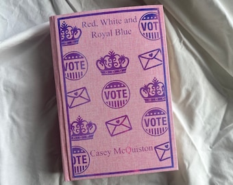 May include: Pink book cover with the title "Red, White and Royal Blue" and the author's name "Casey McQuiston". The cover features a repeating pattern of crowns, envelopes, and the word "VOTE" in a purple and blue color.