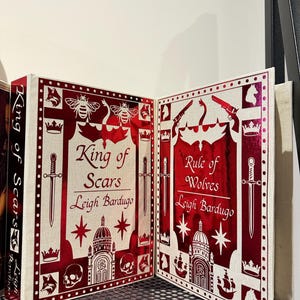 Puede incluir: Dos libros de tapa dura con cubiertas rojas y blancas. El libro de la izquierda se titula "King of Scars" de Leigh Bardugo. El libro de la derecha se titula "Rule of Wolves" de Leigh Bardugo.