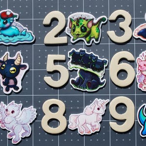 Puede incluir: Una colección de pegatinas con los números del 1 al 9, cada uno emparejado con una criatura de dibujos animados caprichosa. Las criaturas incluyen un monstruo marino, un dragón, una polilla, una cabra, un perro de tres cabezas y un unicornio. Los números son de color beige claro.