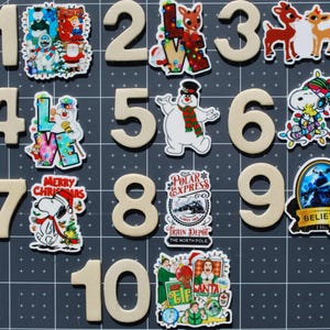 May include: A collection of beige wooden numbers 1 through 10, each paired with a colorful Christmas-themed sticker. Stickers feature snowmen, Snoopy, reindeer, and the words "Believe" and "Polar Express".