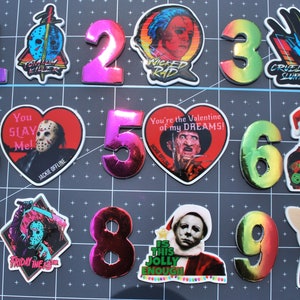 May include: Nine colorful, holographic, vinyl stickers featuring horror movie characters and themes. The stickers are numbered 1 through 9. The stickers feature characters from the movies Friday the 13th, Halloween, A Nightmare on Elm Street, and more.