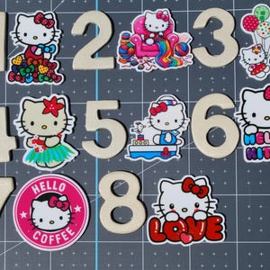 May include: A collection of wooden numbers 1 through 8, paired with colorful Hello Kitty character stickers. The stickers feature Hello Kitty in various poses, including holding balloons, on a boat, and with the word "LOVE". The "Hello Coffee" sticker is also included.