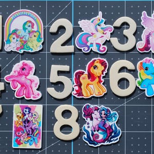 Op de afbeelding: Een verzameling My Little Pony-thema versieringen, inclusief de nummers 1-8 en verschillende pony-karakterontwerpen. De pony's hebben heldere kleuren en zijn geplaatst tegen een rasterachtergrond.
