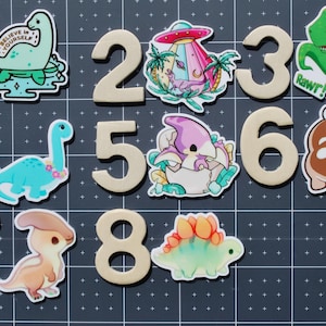 May include: A collection of number-shaped wooden cutouts with dinosaur-themed stickers. The stickers feature various cartoon dinosaurs, including a T-Rex, a Brontosaurus, and a Stegosaurus, along with an alien spaceship and the text "Believe in Yourself".