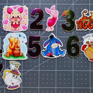 Pode incluir: Uma coleção de adesivos coloridos e números metálicos. Os adesivos apresentam personagens de desenhos animados como Ursinho Pooh, Leitão, Tigrão e Eeyore. Os números de 1 a 7 são exibidos, com um número 3 dourado e um adesivo que diz "lookin like a snack".