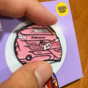 Puede incluir: Un pin de esmalte rosa brillante con forma de casco de carreras con el número 44, el texto UniCredit y el texto Boxboxnow impresos en el lateral. El pin está sujeto a una tarjeta de soporte de color morado claro con un logotipo circular amarillo.