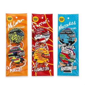 May include: Three vertical retail packages containing circular button pins featuring Formula 1 driver helmet designs. The orange pack shows Lando Norris and Oscar Piastri, the red pack shows Charles Leclerc and Lewis Hamilton, and the blue pack shows George Russell and Kimi Antonelli.