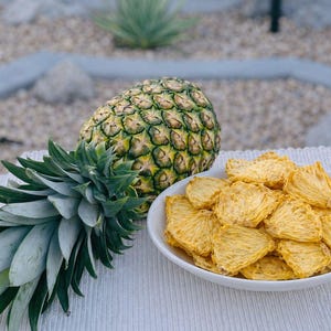 May include: A whole pineapple with green and yellow skin, next to a white bowl filled with dried pineapple slices. The pineapple has a green leafy top. The bowl is on a white surface.