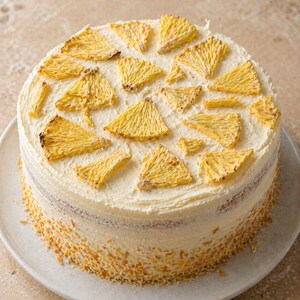 May include: A round cake with white frosting and yellow pineapple slices on top. The cake sits on a white plate and has a layer of yellow crumbs around the bottom. The cake is a dessert.