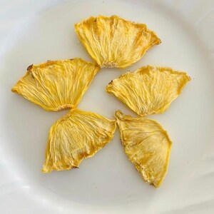 May include: Five dried pineapple slices arranged on a white plate. The pineapple slices are a golden yellow color with a textured, fibrous appearance. The plate is white and has a simple, elegant design. The image is well-lit and the focus is sharp.