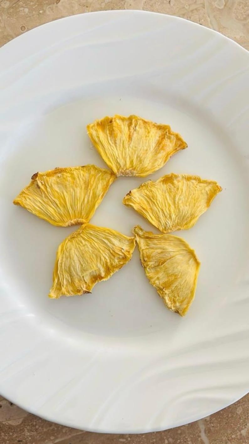 May include: Five dried pineapple slices arranged on a white plate. The pineapple slices are a golden yellow color with a textured, fibrous appearance. The plate is white and has a simple, elegant design. The image is well-lit and the focus is sharp.