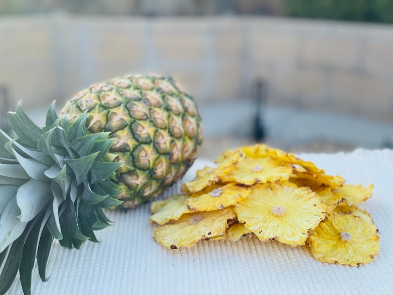 May include: A whole pineapple with green leaves and a textured, green and yellow rind sits next to a pile of dried pineapple slices. The slices are yellow and circular, with a visible core.
