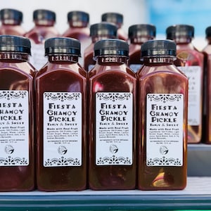 May include: Bottles of Fiesta Chamoy Pickle, a tangy and sweet product. The bottles are filled with a dark red liquid and have black caps. Each bottle has a white label with the product name and ingredients.
