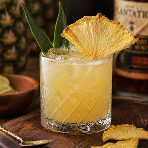 May include: A refreshing cocktail in a diamond-patterned glass, garnished with a dried pineapple slice and green leaves. The drink is set on a wooden board with a gold spoon and pineapple slices. A bottle of Plantation rum is in the background.
