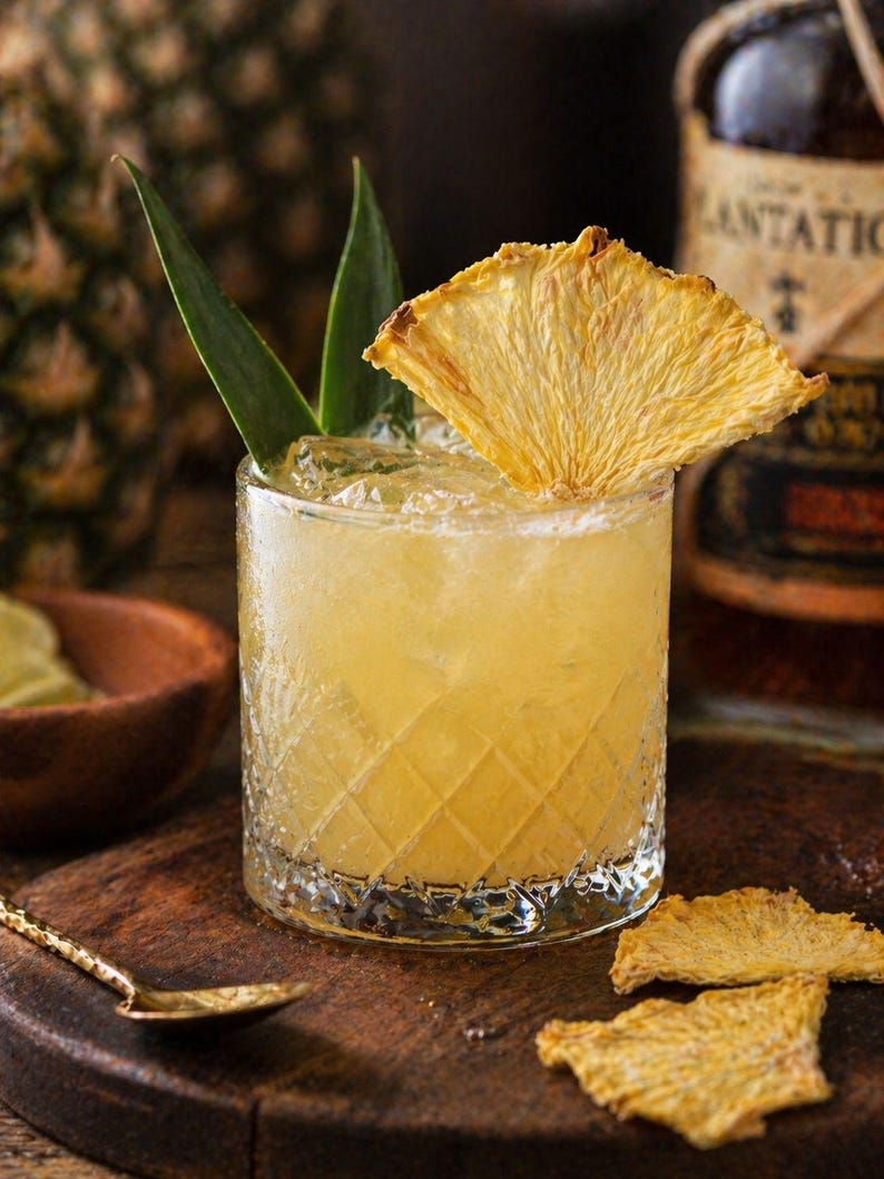 May include: A refreshing cocktail in a diamond-patterned glass, garnished with a dried pineapple slice and green leaves. The drink is set on a wooden board with a gold spoon and pineapple slices. A bottle of Plantation rum is in the background.