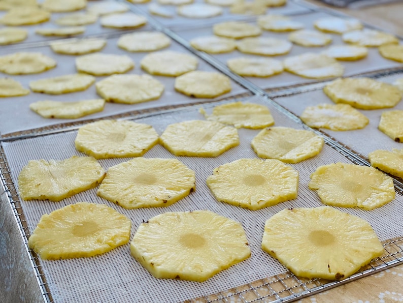 May include: Overhead shot of multiple trays filled with sliced pineapple rings. The pineapple slices are light yellow with a circular pattern and are arranged on a white mesh surface. The trays are on a light-colored surface.