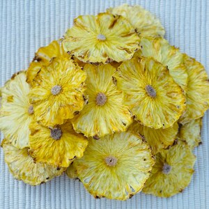 May include: A pile of dried pineapple slices, showcasing a vibrant yellow color with a slightly darker, textured edge. The slices are arranged in a circular pattern, revealing the natural floral design of the fruit. The background is a light, textured surface.