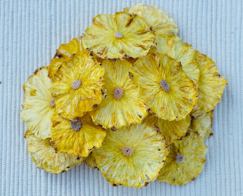 May include: A pile of dried pineapple slices, showcasing a vibrant yellow color with a slightly darker, textured edge. The slices are arranged in a circular pattern, revealing the natural floral design of the fruit. The background is a light, textured surface.