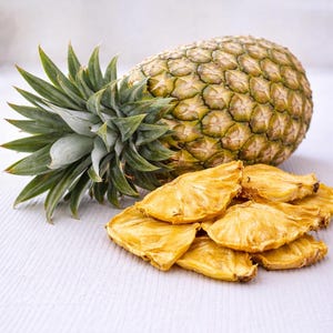 May include: A whole pineapple with green spiky leaves and a textured, segmented exterior. A pile of dried pineapple slices is in front of the fruit. The slices are golden yellow and have a slightly wrinkled appearance.