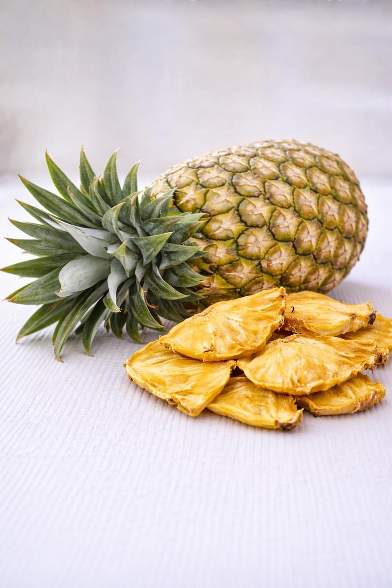 May include: A whole pineapple with green spiky leaves and a textured, segmented exterior. A pile of dried pineapple slices is in front of the fruit. The slices are golden yellow and have a slightly wrinkled appearance.
