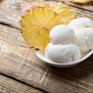 May include: A white, oval-shaped bowl filled with scoops of vanilla ice cream, alongside slices of dried pineapple. The ice cream is stacked, and the pineapple slices are a golden yellow color. The scene is set on a rustic wooden surface.