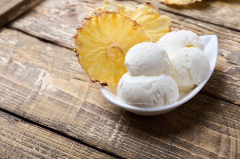 May include: A white, oval-shaped bowl filled with scoops of vanilla ice cream, alongside slices of dried pineapple. The ice cream is stacked, and the pineapple slices are a golden yellow color. The scene is set on a rustic wooden surface.