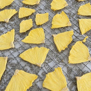 May include: Overhead shot of dried pineapple slices arranged on a metal cooling rack. The slices are a bright yellow color and have a textured, fibrous appearance. The rack sits on a patterned surface.