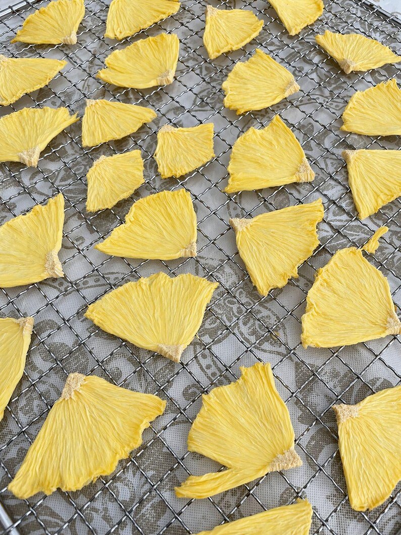 May include: Overhead shot of dried pineapple slices arranged on a metal cooling rack. The slices are a bright yellow color and have a textured, fibrous appearance. The rack sits on a patterned surface.