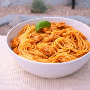 May include: A white bowl filled with spaghetti and chicken in a tomato-based sauce, garnished with a fresh basil leaf. The pasta is arranged in a circular pattern, creating a visually appealing dish.