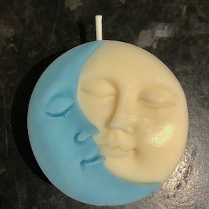 K&ouml;nnte beinhalten: Eine blaue und wei&szlig;e Kerze in Form einer Mondsichel mit einem Gesicht. Der Mond hat geschlossene Augen und einen l&auml;chelnden Mund.