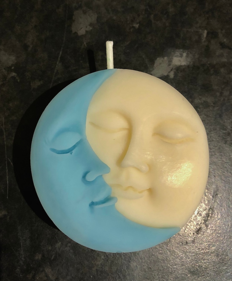 K&ouml;nnte beinhalten: Eine blaue und wei&szlig;e Kerze in Form einer Mondsichel mit einem Gesicht. Der Mond hat geschlossene Augen und einen l&auml;chelnden Mund.