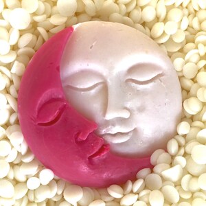 K&ouml;nnte beinhalten: Eine rosa und wei&szlig;e, sichelf&ouml;rmige Seife mit einem Gesicht darauf. Die Seife ist von wei&szlig;en Wachsflocken umgeben.