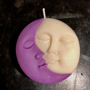 K&ouml;nnte beinhalten: Eine violette und wei&szlig;e Kerze in Form einer Mondsichel und eines Gesichts. Der Mond befindet sich auf der linken Seite der Kerze und das Gesicht auf der rechten Seite. Das Gesicht hat geschlossene Augen und einen l&auml;chelnden Mund.
